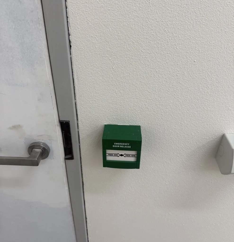 Green break-glass emergency exit release button beside a secured door